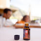A small amber glass doTERRA essential oil bottle with its black cap removed and placed beside it on a light tabletop. The background is heavily blurred, showing an outdoor patio setting with people seated in the distance. doTERRA Endless Oil Blend