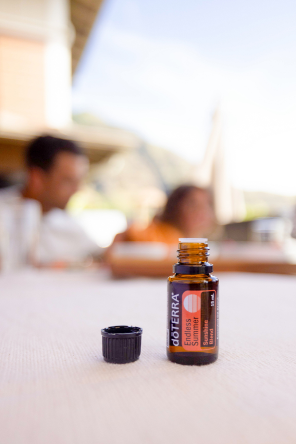 A small amber glass doTERRA essential oil bottle with its black cap removed and placed beside it on a light tabletop. The background is heavily blurred, showing an outdoor patio setting with people seated in the distance. doTERRA Endless Oil Blend
