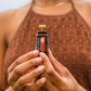 A person in a brown patterned halter top holds a small amber glass bottle of doTERRA essential oil against a soft blurred light gray background. doTERRA Endless Oil Blend