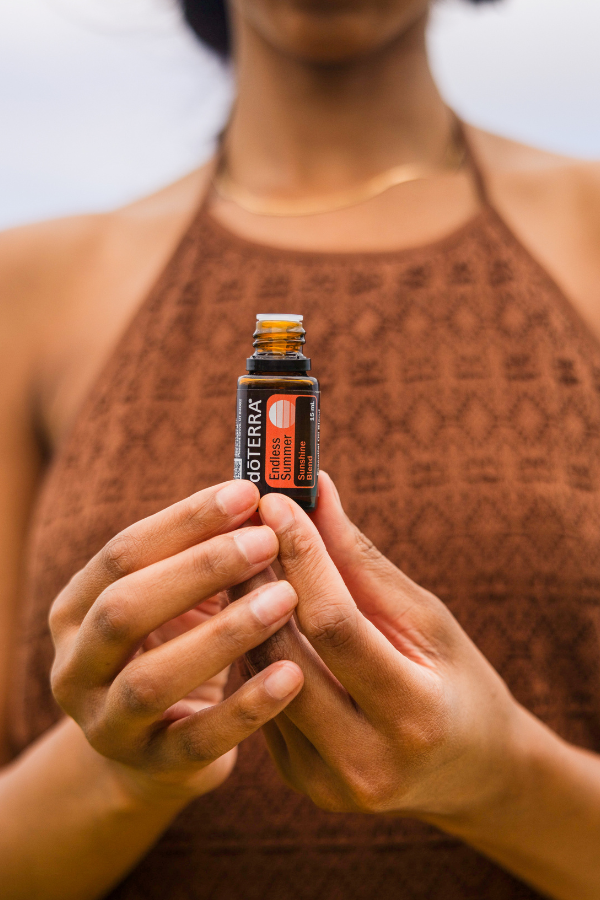 A person in a brown patterned halter top holds a small amber glass bottle of doTERRA essential oil against a soft blurred light gray background. doTERRA Endless Oil Blend