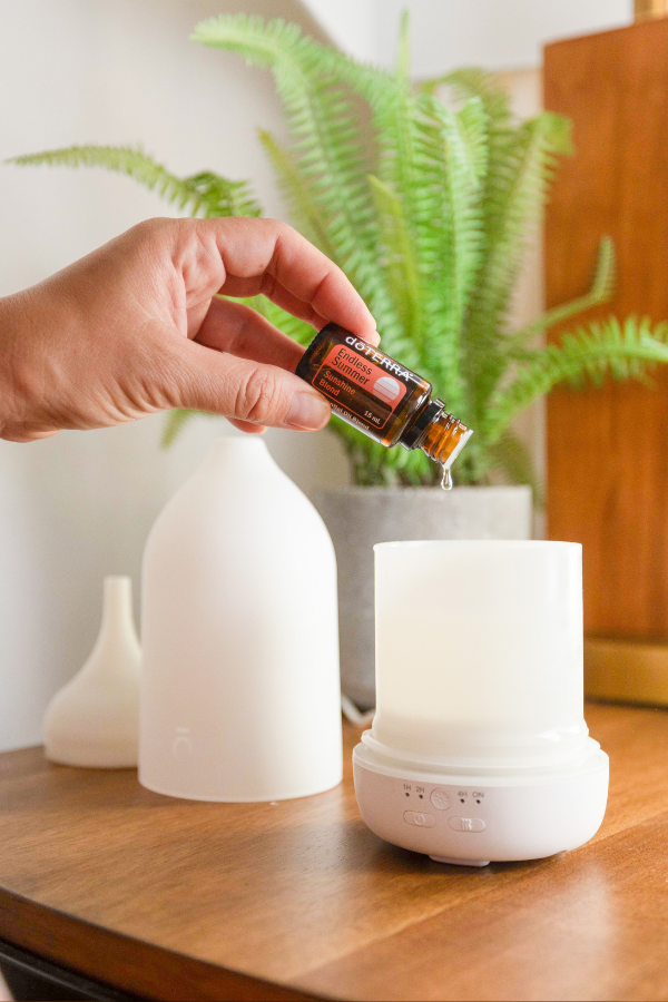 A hand holding a small amber essential oil dropper above a white diffuser on a wooden table, with a larger white diffuser bottle and a green potted fern in the background. doTERRA Endless Oil Blend