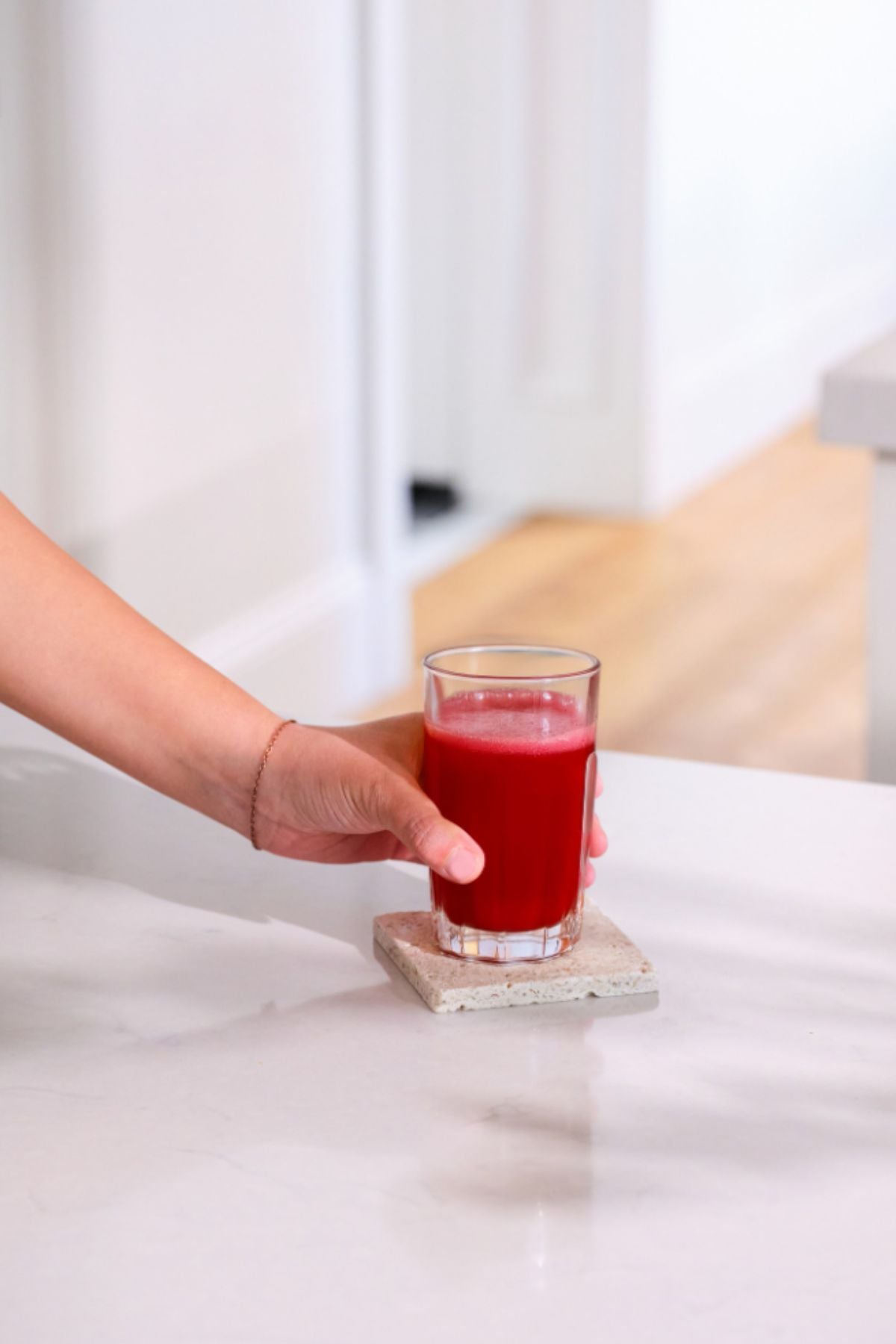 A hand holding a clear glass filled with bright red juice, resting on a square concrete coaster on a white marble countertop in a bright kitchen setting. dōTERRA MetaPWR Recharge Electrolytes (Wild Strawberry)