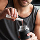 A man in a gray tank top mixes a red liquid into a clear shaker bottle with a black sleeve, holding a white and orange  MicroPlenty packet above the bottle. dōTERRA MetaPWR Recharge Electrolytes (Wild Strawberry)