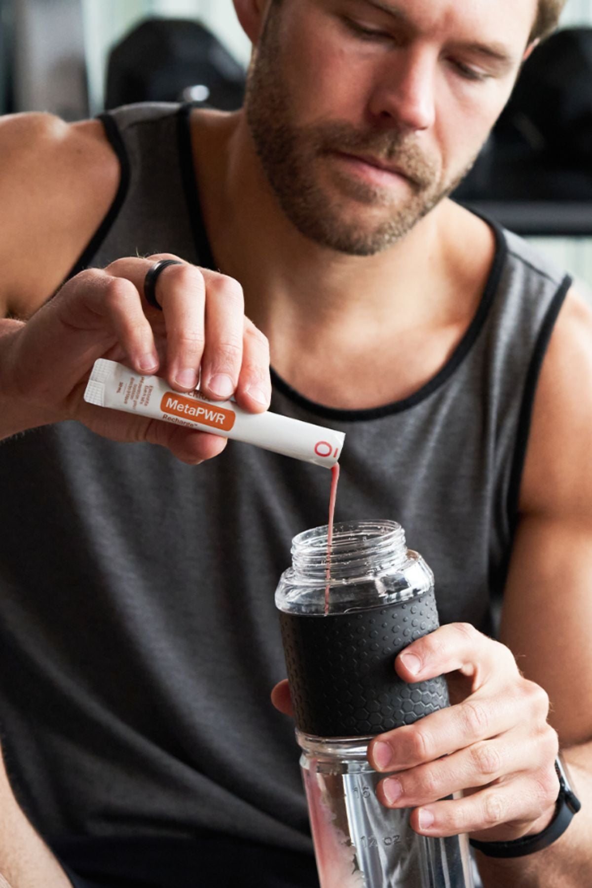 A man in a gray tank top mixes a red liquid into a clear shaker bottle with a black sleeve, holding a white and orange  MicroPlenty packet above the bottle. dōTERRA MetaPWR Recharge Electrolytes (Wild Strawberry)